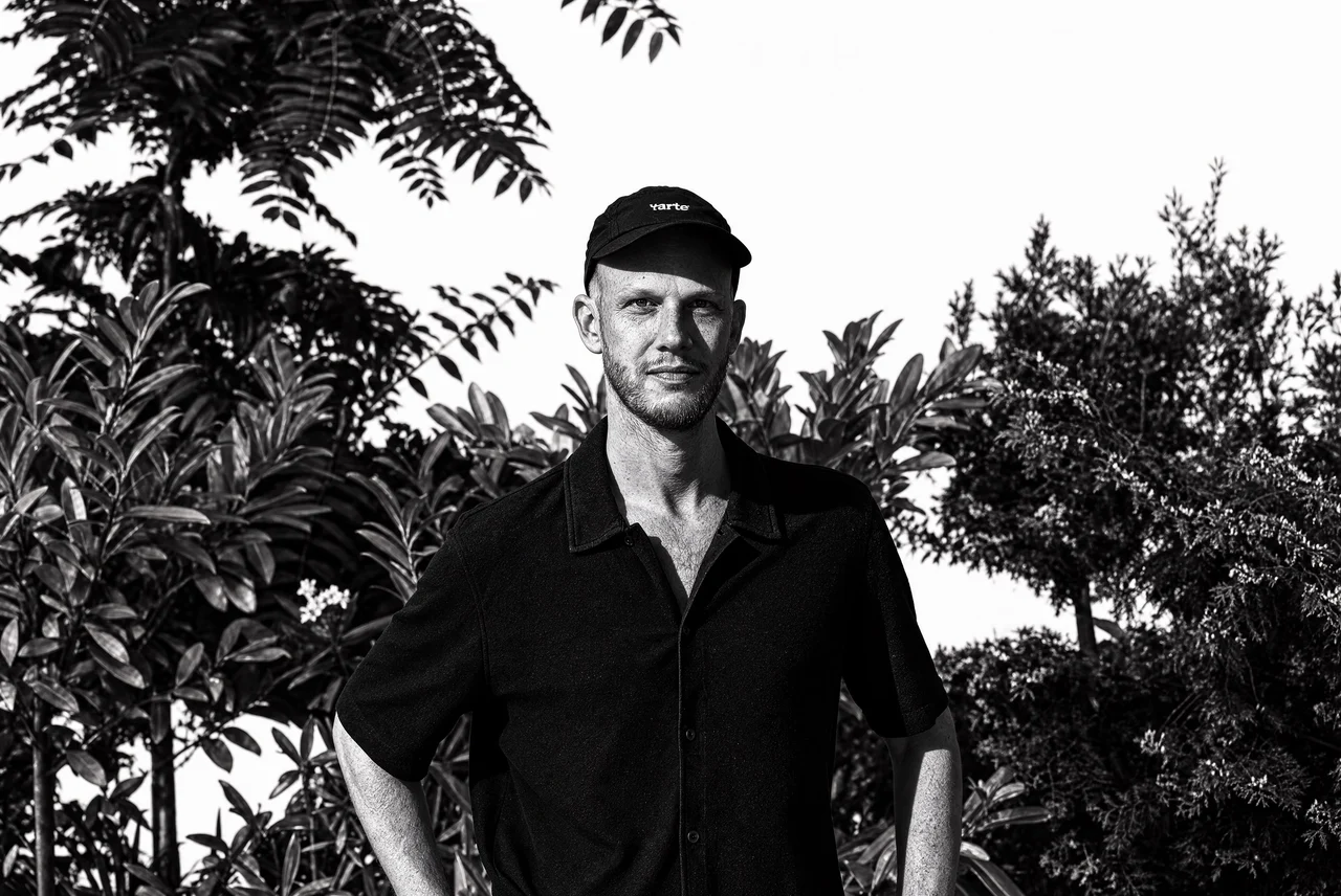 Philipp John in a black shirt and cap in front of trees, black and white